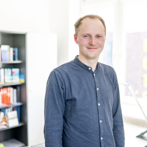 Eine Person mit einem gemusterten blauen Hemd steht in einem hellen, modernen Büro mit Bücherregalen und großen Fenstern im Hintergrund.